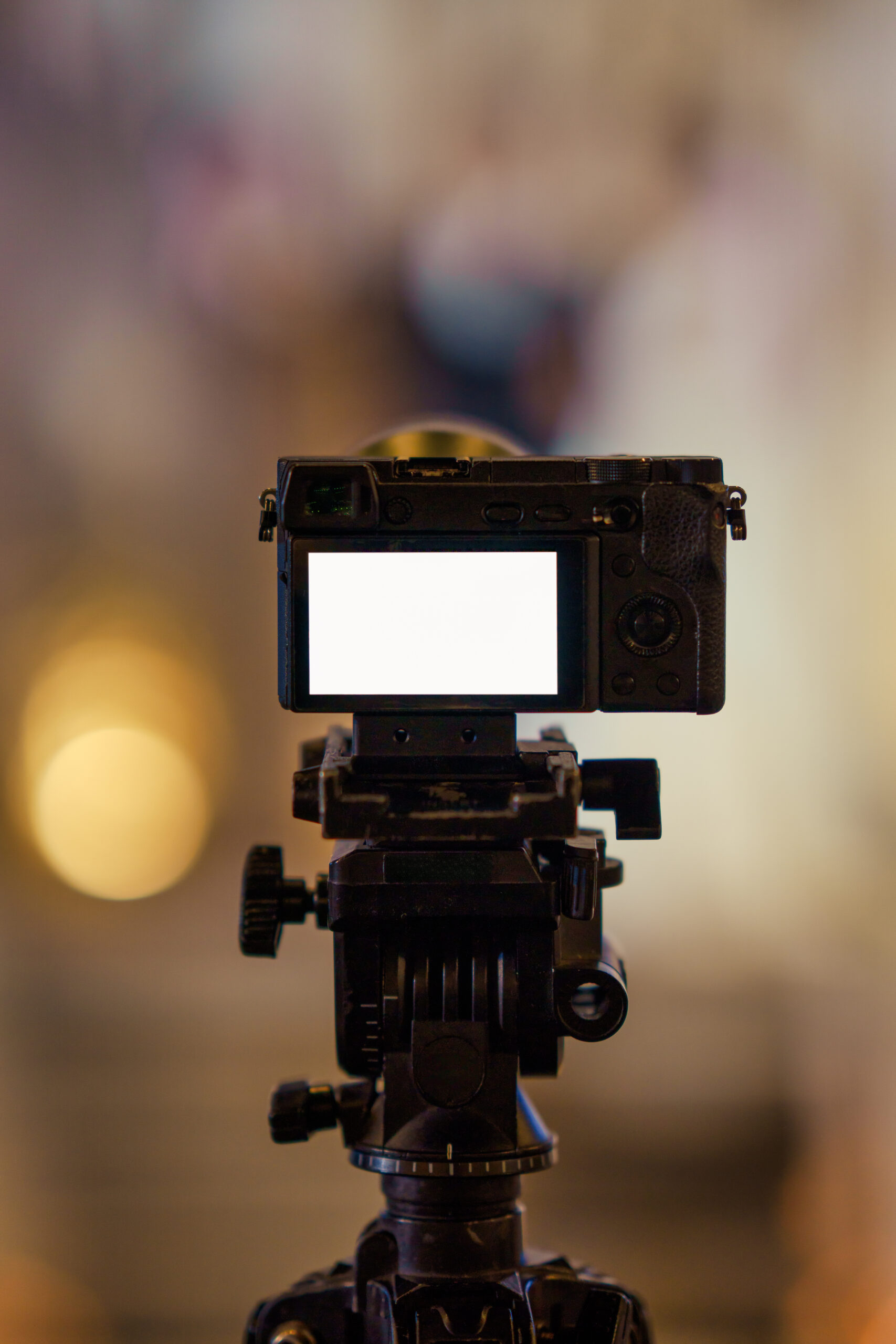 Closeup of professional mirrorless camera filming video on ball head tripod with blank LCD screen on blurred golden background in wedding ceremony